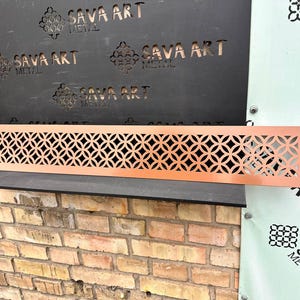 Può includere: Un pannello decorativo in metallo color rame con un motivo geometrico di cerchi interconnessi. Il pannello rettangolare presenta una serie di disegni ritagliati. Lo sfondo include un muro di mattoni e un pannello nero con la scritta "SAVA ART METAL".