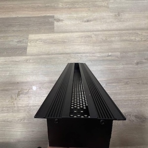 May include: Black metal light fitting with a rectangular shape and a grid pattern. The fitting has a long, narrow design with a series of parallel lines running along the top and bottom.