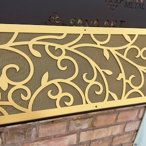 May include: A gold metal decorative vent cover with a floral design. The vent cover is made of metal and has a mesh screen behind the design. The vent cover is mounted on a brick wall.