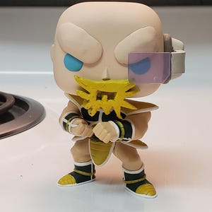 Puede incluir: Una figura de personaje de anime amarillo y negro con cabeza calva, ojos azules y barba amarilla. La figura lleva un atuendo amarillo y negro y tiene una visera morada en el ojo derecho.