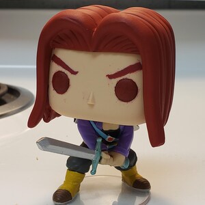 May include: A red-haired anime character figurine holding a silver sword. The figurine is wearing a purple shirt and black pants. The figurine is standing on a clear plastic base.
