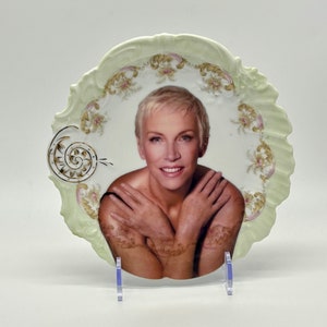 May include: A white porcelain plate with a green and gold floral design. A woman with short blonde hair is pictured in the center of the plate.