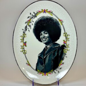 May include: Oval decorative plate featuring a black and white portrait of a smiling person with a large afro. The plate has a floral border with a silver rim. The background is white with a delicate embossed pattern.
