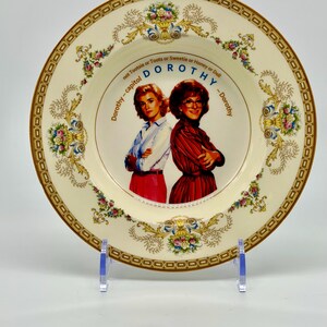 May include: A decorative plate featuring two women with the text "Dorothy" and "Dorothy - capitol DOROTHY - Dorothy". The plate has a cream-colored background with floral and gold accents around the rim and the central image.