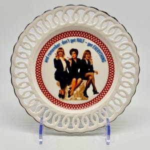 May include: A white decorative plate with a pierced edge and a black rim. The center features an image of three women and the text "and remember, don't get HALF - get EVERYTHING". The plate is displayed on a clear stand.