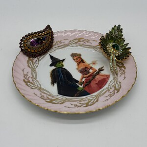 May include: A decorative pink plate with a gold rim and a Wizard of Oz theme. The plate features a picture of the Wicked Witch of the West and Glinda the Good Witch. Two decorative brooches are on the plate, one is a teardrop shape with a purple stone.
