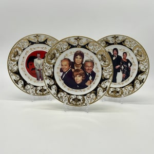 May include: Three white ceramic plates with gold and black trim. Each plate features a different image of a group of people. The plates are on a white surface.