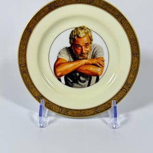 May include: A round ceramic decorative plate featuring a central portrait of a person with gray hair and a beard, wearing a gray t-shirt. The plate has a cream-colored center and a wide, ornate gold and black border with a detailed floral pattern.