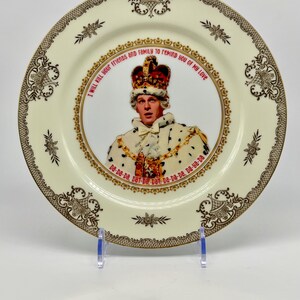 May include: A decorative ceramic plate with a portrait of a man wearing a crown and royal attire. The plate has a cream-colored background with a gold and brown ornate border. Text on the plate reads "I will kill your friends and family to remind you of my love."