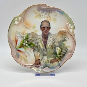 May include: Decorative plate featuring a portrait of a person wearing sunglasses and a white suit. The plate has a scalloped edge with a gold trim and a floral design in pastel colors. The plate is on a clear stand.