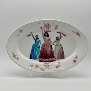 May include: Oval white ceramic platter featuring a design of three women in period dresses, labeled "Eliza," "Angelica," and "Peggy." The text "The Schuyler Sisters" is below the figures. Pink floral accents and a decorative border complete the design.
