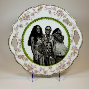 May include: A decorative serving tray with a scalloped edge, featuring a black and white image of three people. The tray has a green and gold border, floral accents, and gold handles. The tray is displayed on a clear stand.