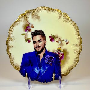 May include: Decorative plate with a portrait of a person in a blue suit, adorned with a purple flower. The plate has a cream-colored background with floral accents and a gold-colored, scalloped edge. The plate is displayed on a clear stand.