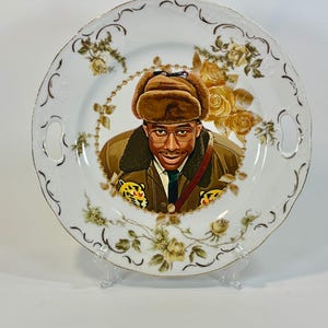 May include: A white decorative plate with a gold rim and handles, featuring a portrait of a person wearing a fur hat and glasses. The plate is adorned with floral and gold accents, and a tiger patch.