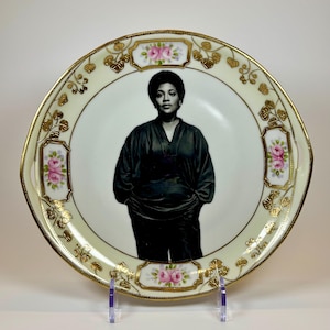May include: A decorative plate featuring a black and white portrait of a person. The plate has a cream-colored border with gold accents and pink rose details. The plate is displayed on a clear stand.