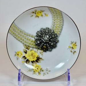 May include: A white decorative plate with a silver rim, featuring a design of a pearl necklace and a black and silver brooch. Yellow roses are scattered around the plate. The plate is displayed on a clear plastic stand.