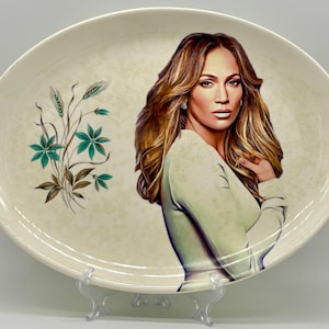 May include: A white oval ceramic plate with a floral design and a portrait of a woman with long brown hair. The woman is wearing a white shirt and has a neutral expression.