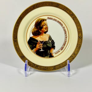 May include: A round decorative ceramic plate with a cream-colored center and a wide, ornate gold-patterned border. The center features an illustration of a person in a dark dress holding a cocktail glass, with the text: Fasten your seatbelts, it's going to be a bumpy night. - Margo Channing.