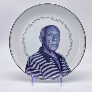 May include: A white ceramic plate with a silver rim features a black and white portrait of a man with a striped shirt. The plate has a blue and silver floral design around the edge.