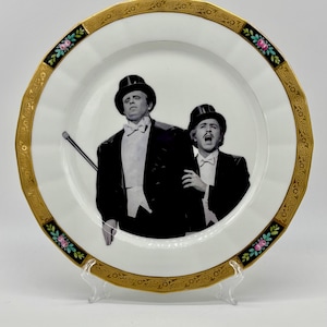 May include: A decorative white plate with a gold rim and a black and pink floral design. The plate features a black and white image of two men in tuxedos and top hats. The plate is displayed on a clear stand.