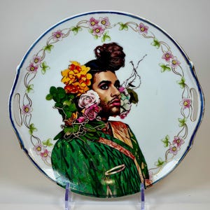 May include: A decorative ceramic serving platter featuring a portrait of a person adorned with flowers and greenery. The person is wearing a green and gold patterned garment. The platter has a white background with a floral border and gold trim.