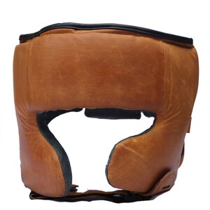 May include: A brown leather boxing headguard with black trim and a padded interior. The headguard has a strap that goes under the chin.