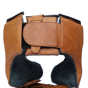 May include: A brown leather boxing headguard with black padding and adjustable straps. The headguard has a velcro closure at the back.