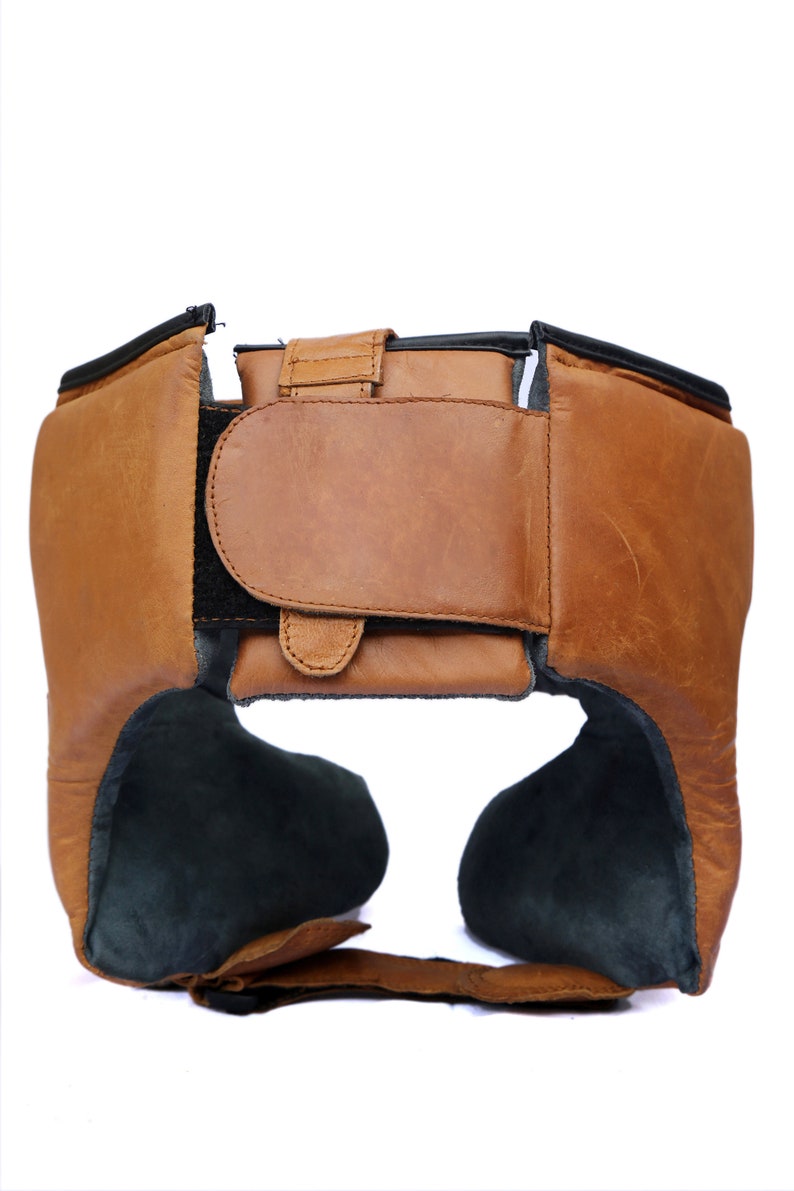 May include: A brown leather boxing headguard with black padding and adjustable straps. The headguard has a velcro closure at the back.