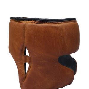 May include: A brown leather boxing headguard with black trim. The headguard has a padded top and a chin strap.