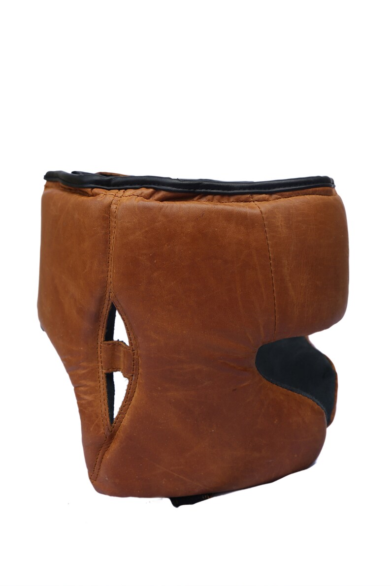May include: A brown leather boxing headguard with black trim. The headguard has a padded top and a chin strap.
