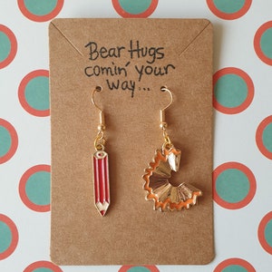 May include: A pair of gold earrings on a brown card with the text "Bear Hugs comin' your way..." One earring is a red pencil and the other is a gold pencil sharpener.