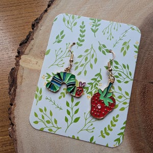 May include: A pair of gold-toned dangle earrings. One earring is a colorful caterpillar, the other a red strawberry with green leaves. The earrings are displayed on a white card with green leaf patterns, set on a wooden surface.