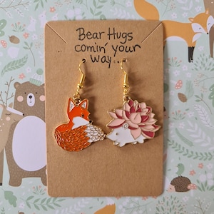 May include: A pair of gold dangle earrings featuring a fox and a hedgehog charm. The fox is orange and white with a red nose. The hedgehog is white with a pink and red flower on its back. The earrings are displayed on a brown card with the text "Bear Hugs comin' your way..."