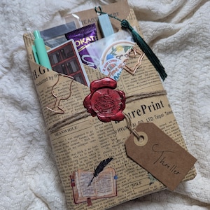 May include: A wrapped gift package with a vintage newspaper print. It's sealed with a red wax stamp and tied with twine. A tag reads "Thriller." Inside are pens, stickers, and a chocolate bar. A green tassel hangs from the top.