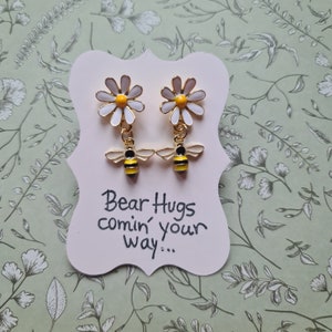 May include: A pair of gold earrings with a white daisy and a yellow and black bee charm. The earrings are displayed on a white card with the text "Bear Hugs comin' your way..."