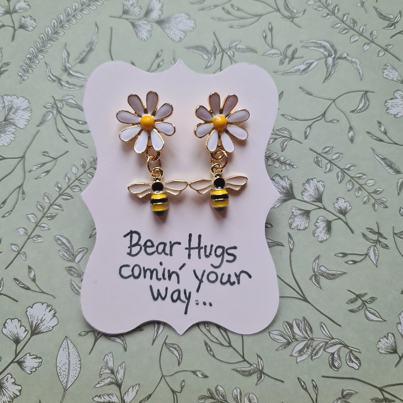 May include: A pair of gold earrings with a white daisy and a yellow and black bee charm. The earrings are displayed on a white card with the text "Bear Hugs comin' your way..."