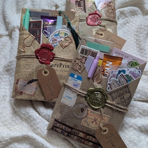 May include: Three wrapped packages with decorative elements. The packages are wrapped in brown paper and tied with twine. Each package has a wax seal and a tag that reads "Thriller", "Romance", and "Fiction". The packages contain stickers, pens, and other small items.