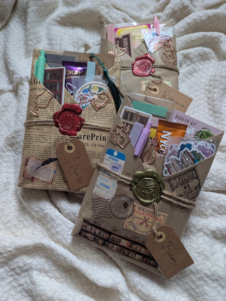 May include: Three wrapped packages with decorative elements. The packages are wrapped in brown paper and tied with twine. Each package has a wax seal and a tag that reads "Thriller", "Romance", and "Fiction". The packages contain stickers, pens, and other small items.