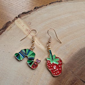 May include: A pair of gold-toned dangle earrings. One earring features a colorful caterpillar design, and the other has a red strawberry with green leaves. The earrings are set against a wooden background.