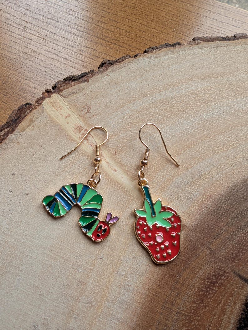 May include: A pair of gold-toned dangle earrings. One earring features a colorful caterpillar design, and the other has a red strawberry with green leaves. The earrings are set against a wooden background.