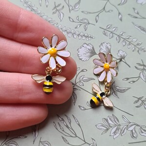 May include: A pair of dangle earrings featuring a white daisy with a yellow center and a gold bee with black and yellow stripes.
