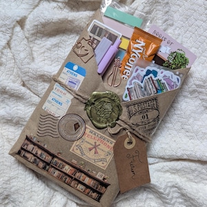 May include: A brown paper package tied with twine, sealed with a wax stamp, and adorned with travel-themed stickers and stamps. The package contains stickers, stationery, and a small snack. A tag reads "Fiction".