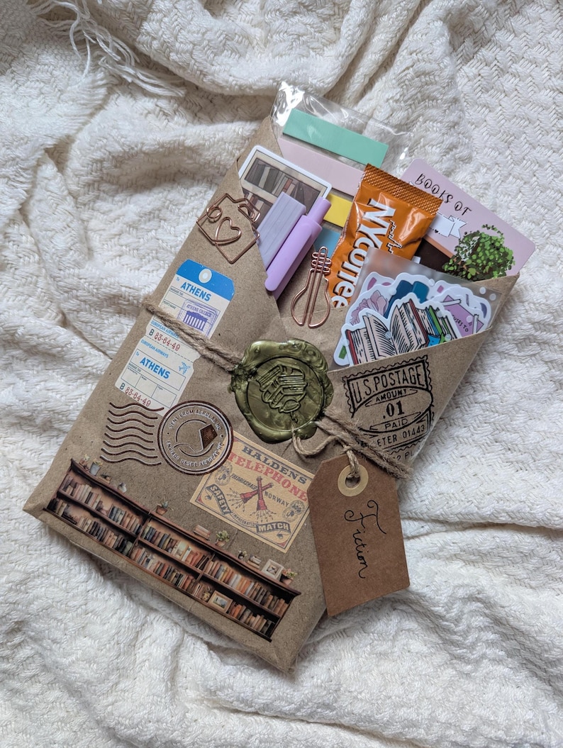 May include: A brown paper package tied with twine, sealed with a wax stamp, and adorned with travel-themed stickers and stamps. The package contains stickers, stationery, and a small snack. A tag reads "Fiction".