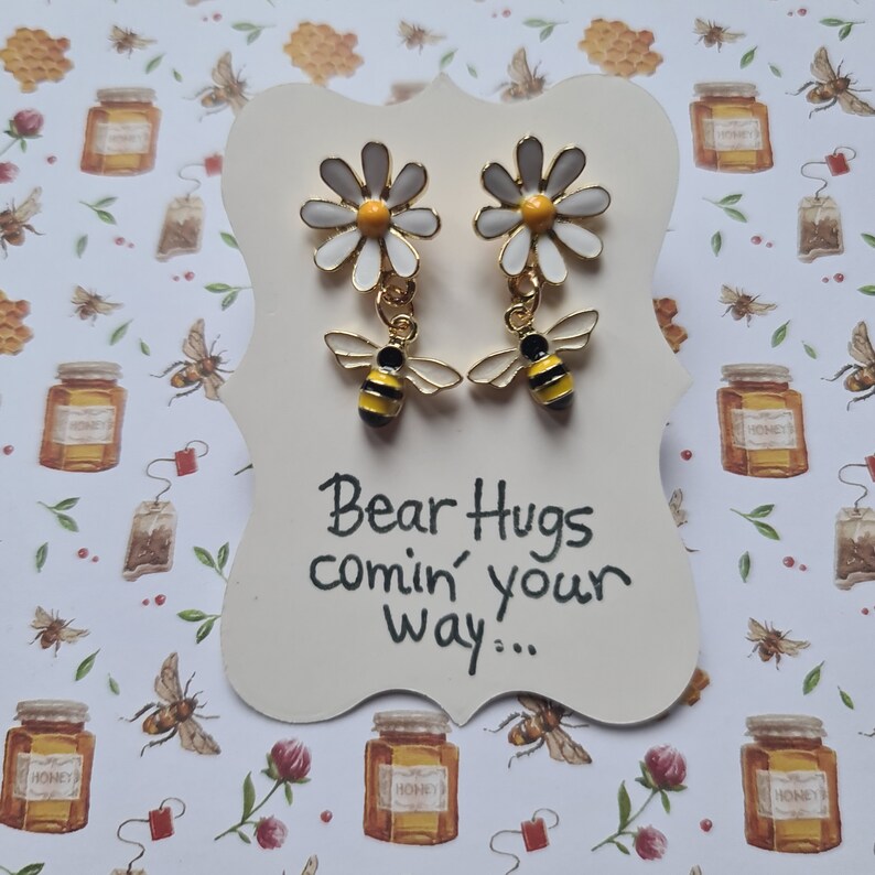 May include: A pair of gold earrings with a white daisy and a yellow and black bee charm. The earrings are on a white card with the text "Bear Hugs comin' your way..."