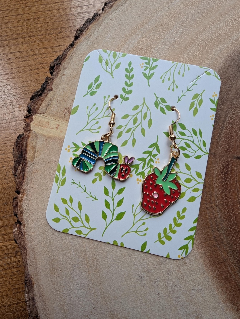 May include: A pair of gold-toned dangle earrings. One earring is a colorful caterpillar, the other a red strawberry with green leaves. The earrings are displayed on a white card with green leaf patterns, set on a wooden surface.