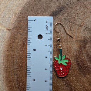 May include: A gold-colored earring with a red strawberry design. The strawberry has green leaves and a gold outline. The earring measures approximately 1.5 inches in length, including the hook.