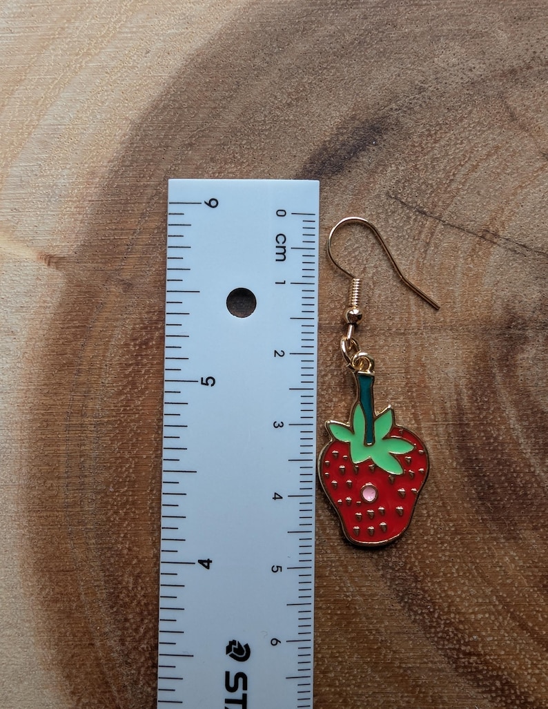 May include: A gold-colored earring with a red strawberry design. The strawberry has green leaves and a gold outline. The earring measures approximately 1.5 inches in length, including the hook.
