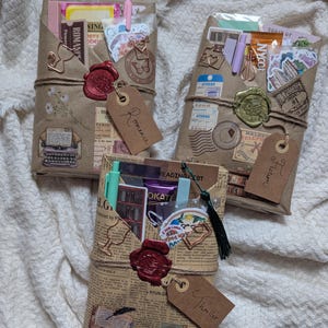 May include: Three wrapped gifts, each with a different theme: Romance, Fiction, and Thriller. The gifts are wrapped in brown paper and newspaper, sealed with wax, and adorned with tags. Each gift contains stickers, pens, and other small items.