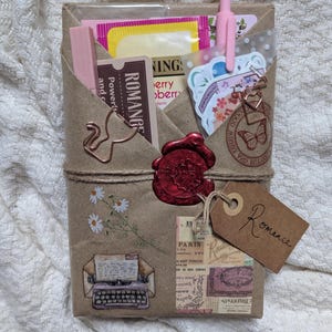 May include: A wrapped gift package with a brown paper exterior, tied with twine. It features decorative stickers, a red wax seal, and a tag that says "Romance". The package contains pink items, tea bags, and stationery.