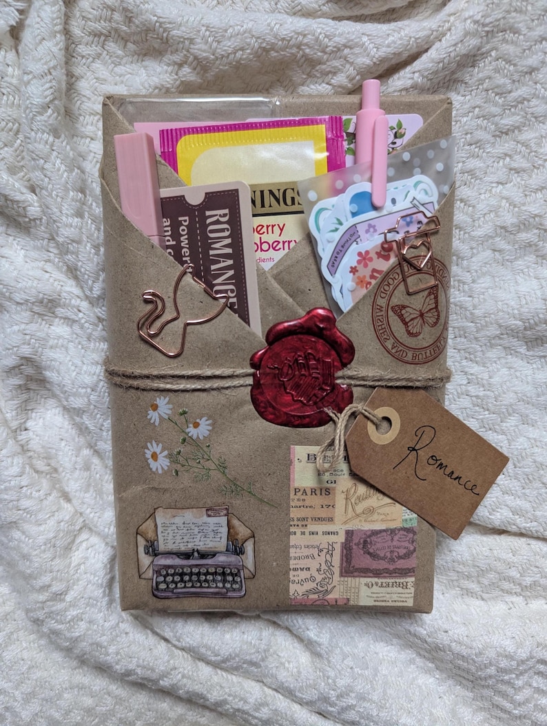 May include: A wrapped gift package with a brown paper exterior, tied with twine. It features decorative stickers, a red wax seal, and a tag that says "Romance". The package contains pink items, tea bags, and stationery.